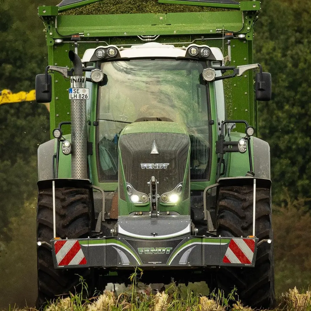 Green tractor with large tires and a Fendt front weight bumper, shown head-on in a field with greenery behind
