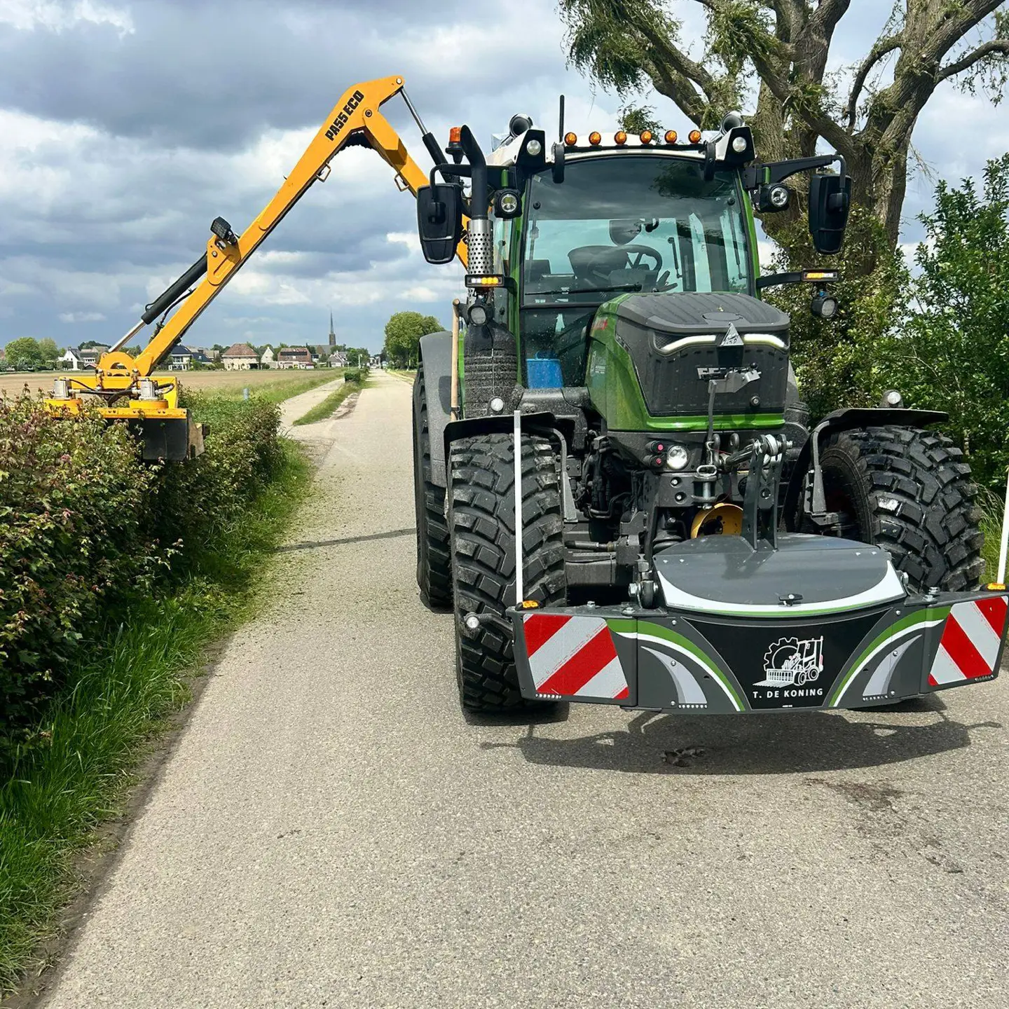 A green tractor with a front weight bumper labeled T. DE KONING is parked on a rural road next to hedges.