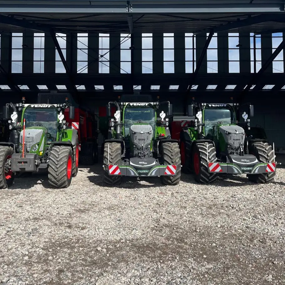Three green tractors with front weight bumpers are parked side by side on gravel under a large metal-roofed structure