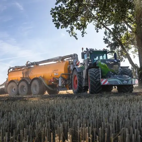 Un tracteur vert avec un grand pare-chocs poids avant tire une remorque-citerne orange à travers un champ récolté
