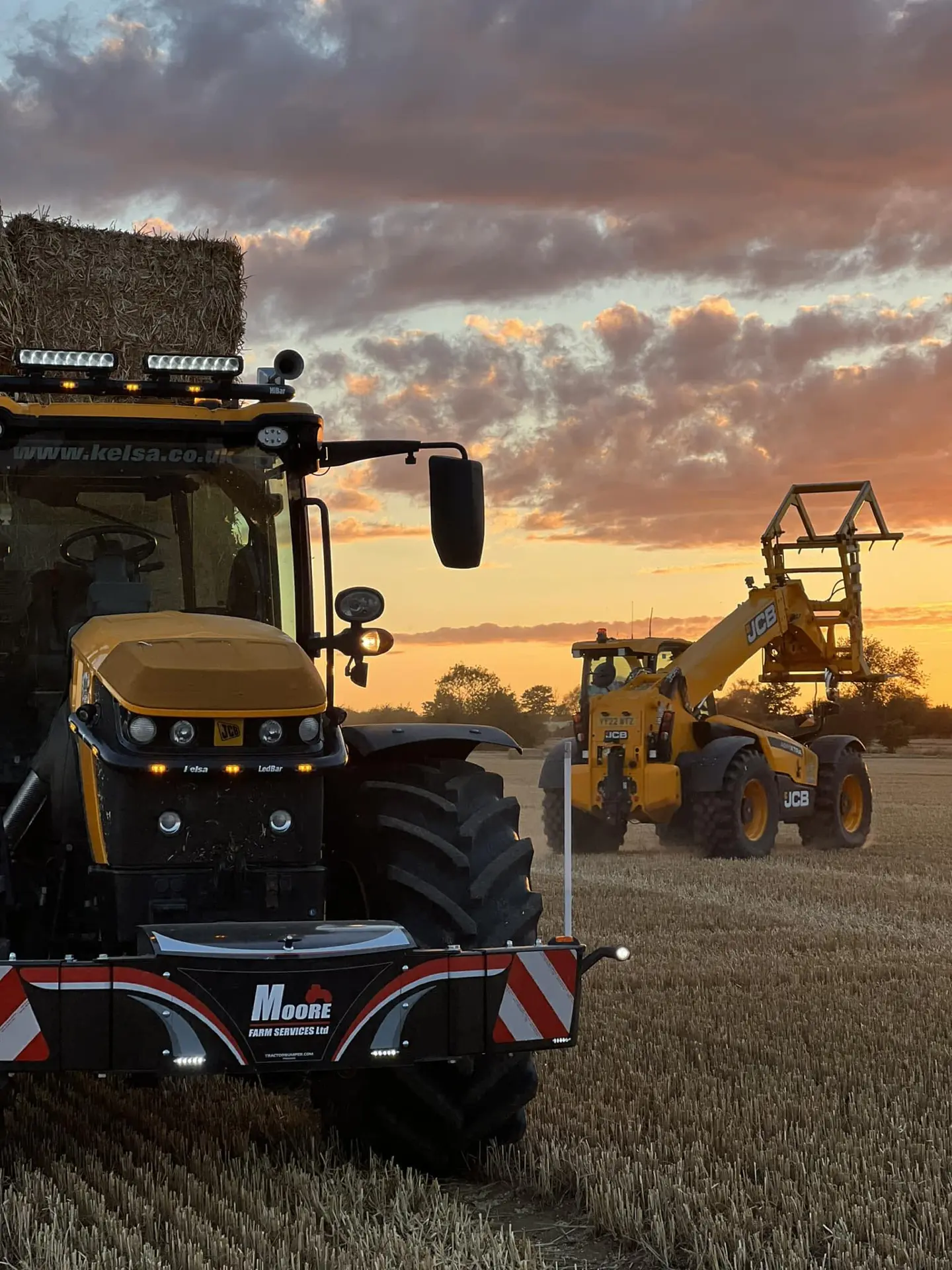 Front view of a tractor in a field at sunset with a front weight bumper and LED lights, carrying hay bales.