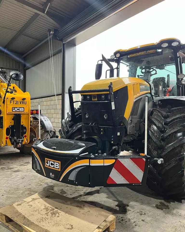 A yellow tractor indoors with a mounted front weight bumper featuring JCB branding and red reflective safety stripes