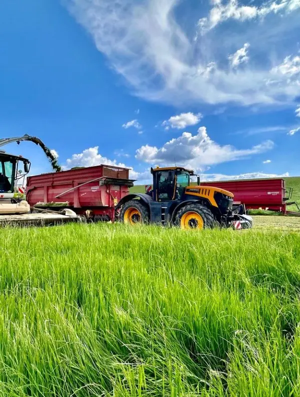 A yellow and black tractor with a front weight bumper is attached to a red trailer in a grassy field under a blue sky