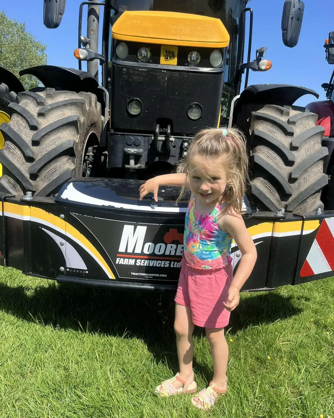 A young girl stands in front of a tractor fitted with a front weight bumper labeled Moore Farm Services Ltd
