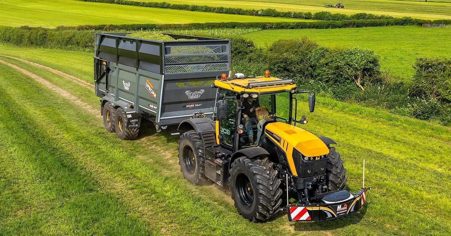 A yellow JCB tractor with a large front weight bumper and LED lights pulls a green trailer on a grassy field