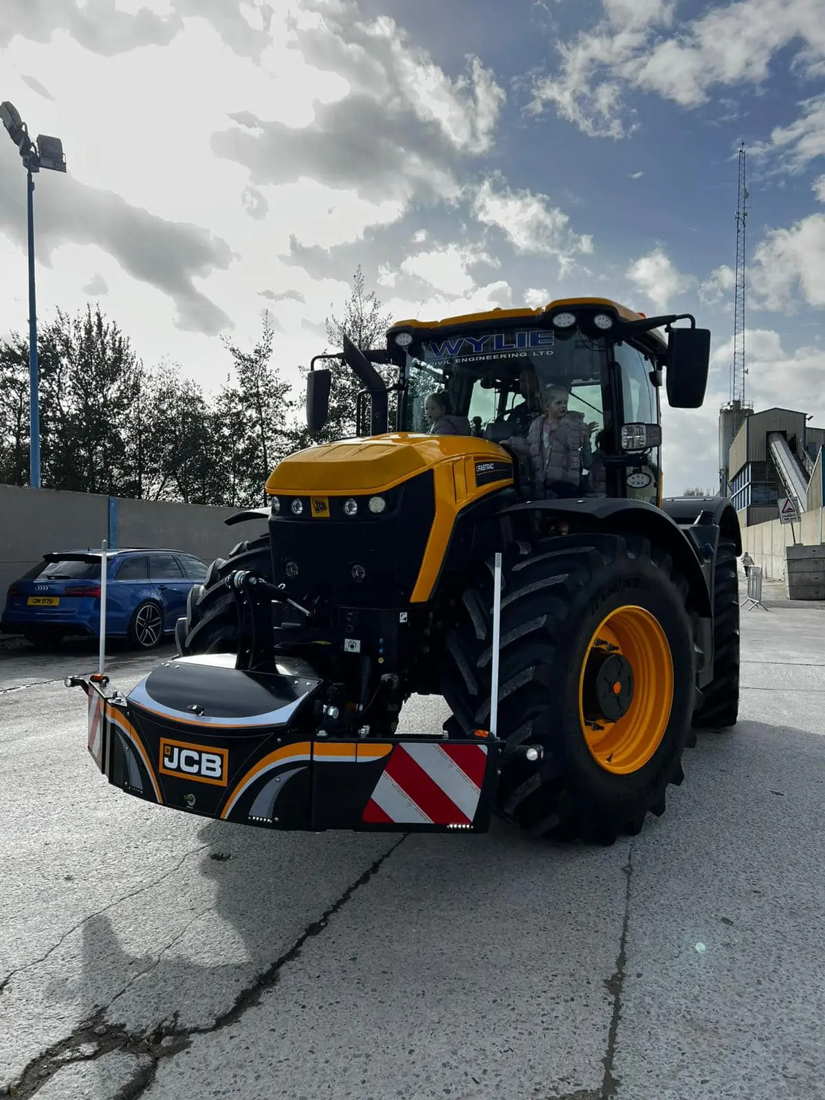Yellow and black tractor with a front weight bumper labeled JCB, parked on concrete with two people in the cab