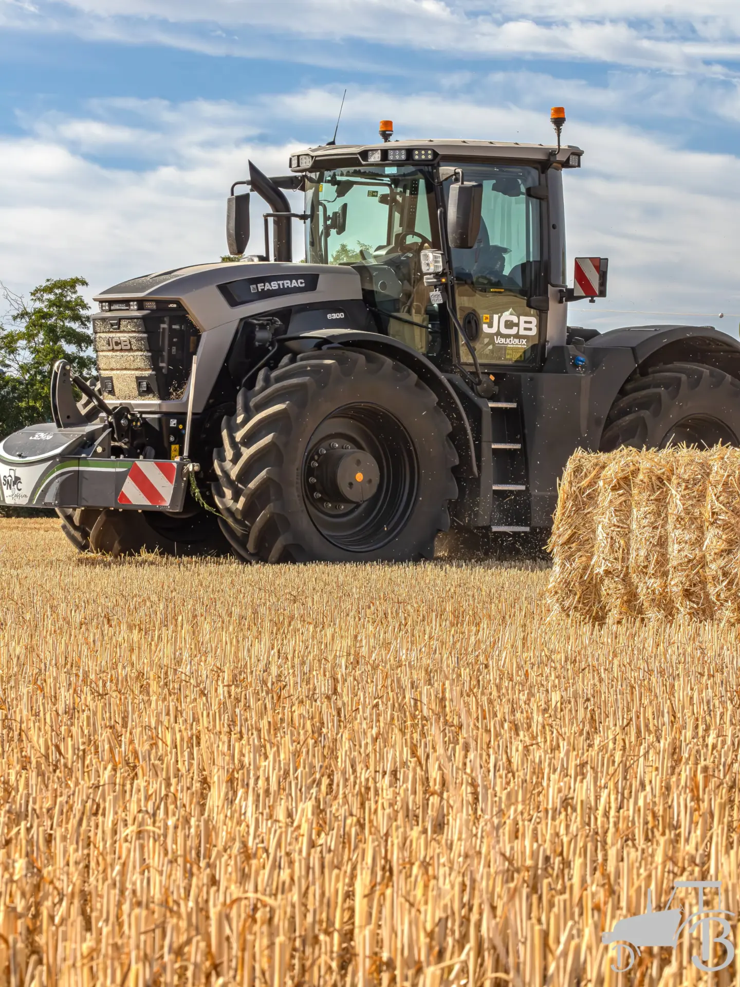A tractor with a front weight bumper and LED lights is working in a harvested field beside a round bale of straw