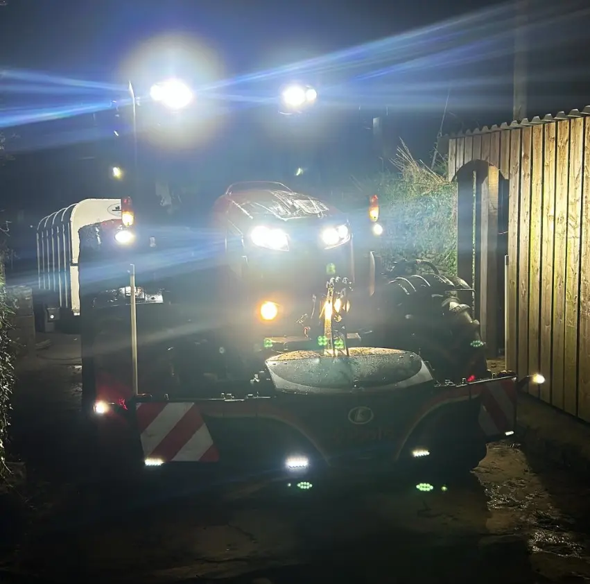 A tractor with illuminated headlights and a mounted front weight bumper is parked on a farm path at night