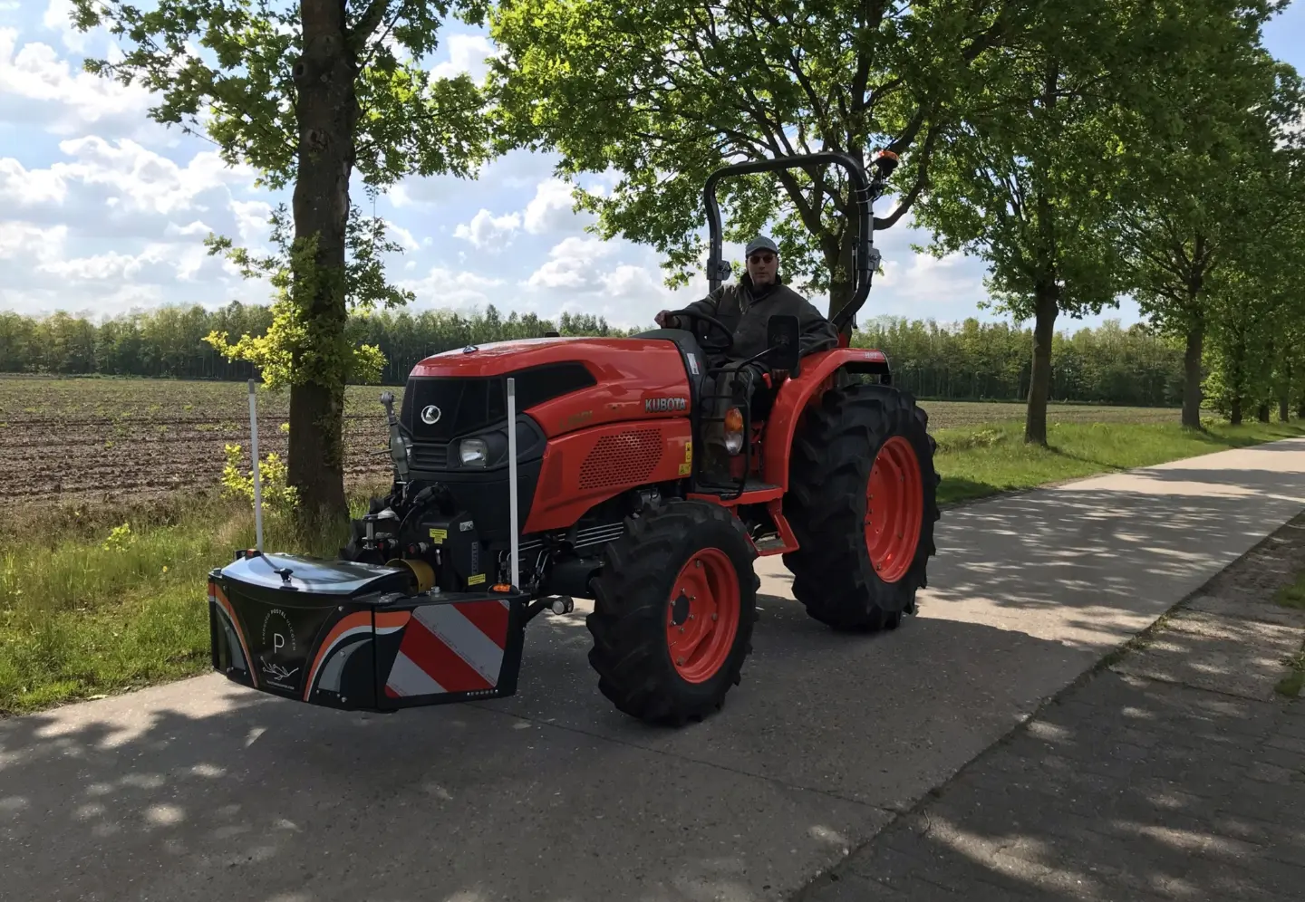 A man drives an orange tractor with a mounted front weight bumper on a paved road beside a field and trees