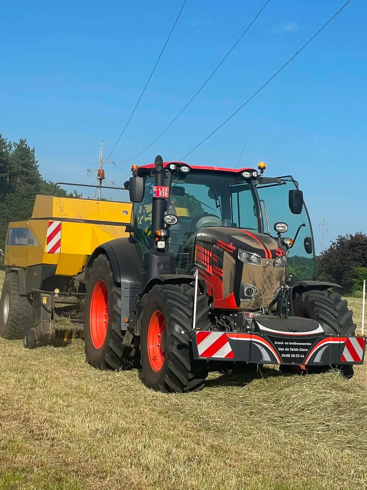 A tractor with a front weight bumper and red reflective stripes is working in a field, attached to a yellow baler.