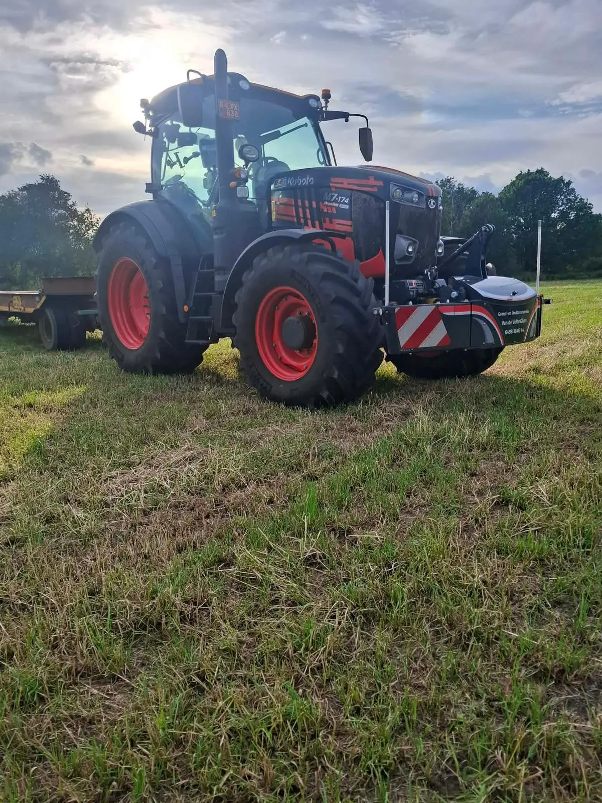 A tractor with a front weight bumper is parked on grass with a trailer attached in the background