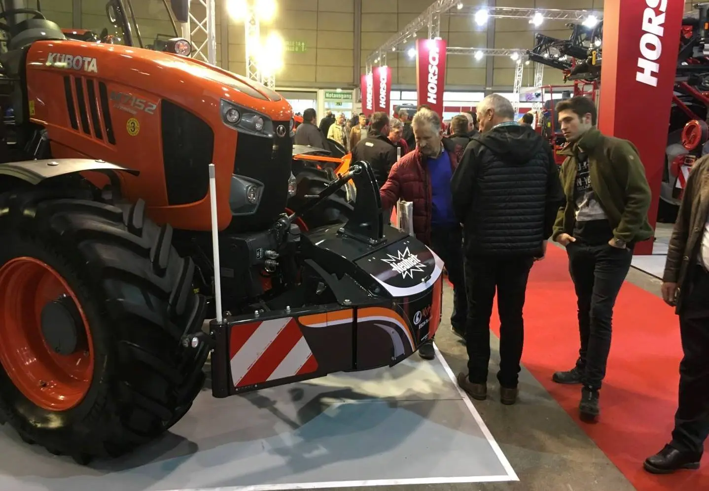 A group of people examine a tractor equipped with a front weight bumper and safety markings at an indoor expo