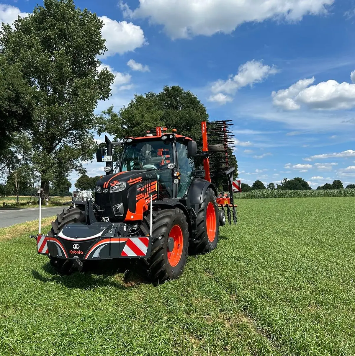 A tractor with a front weight bumper and rear attachment stands on grass near a road under a partly cloudy sky