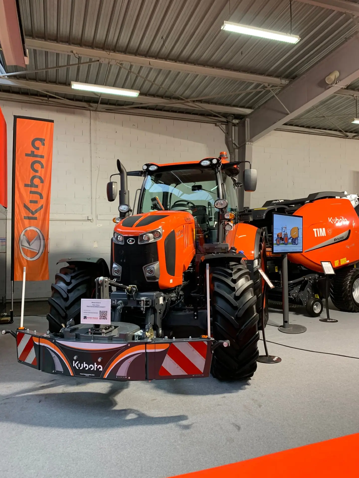 Orange tractor on display indoors with a large front weight bumper labeled Kubota and red-white safety stripes