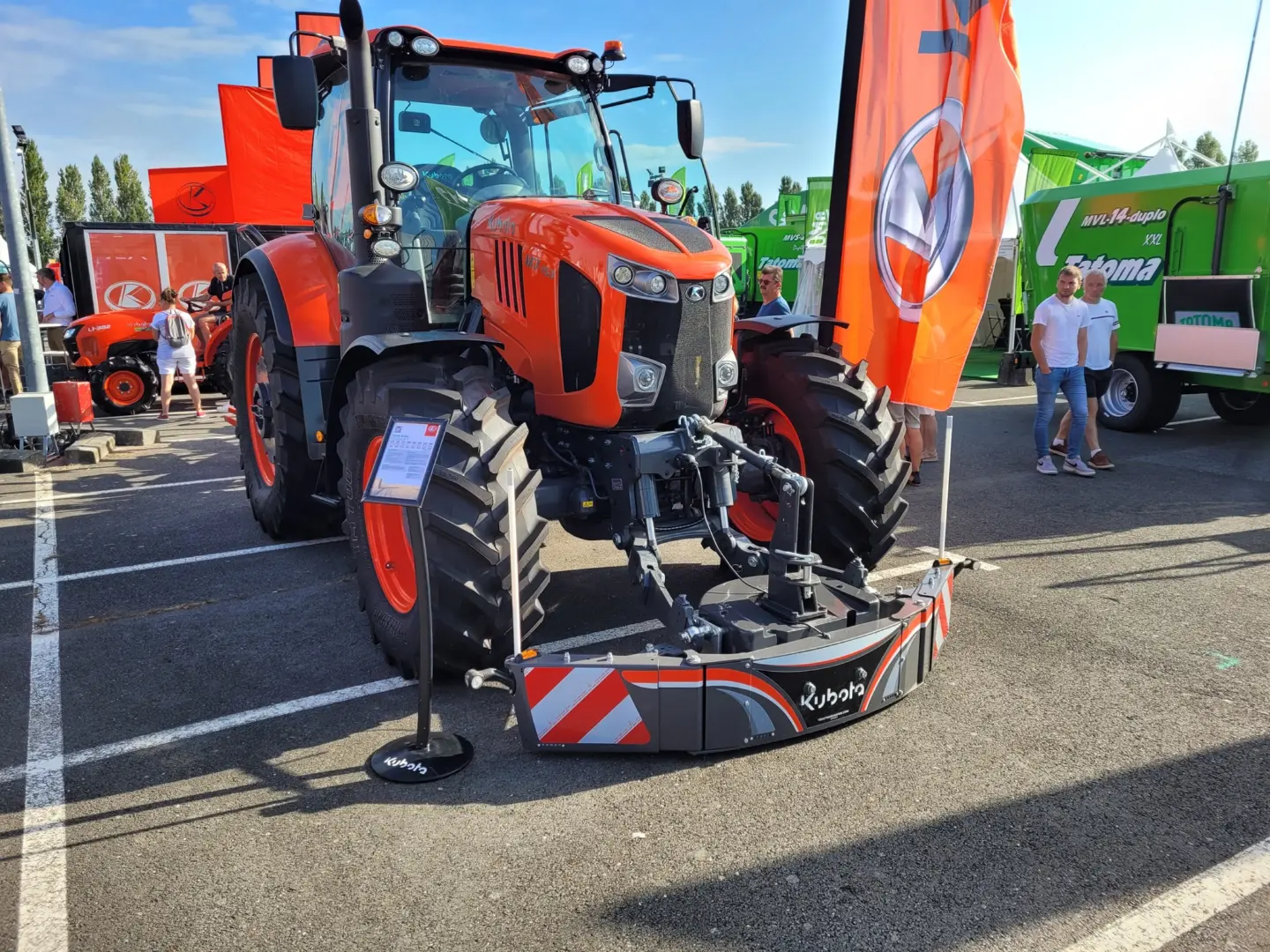 Large orange tractor with a front weight bumper attached, displayed outdoors with people and banners nearby