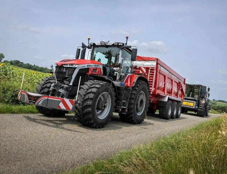 Red tractor with a front weight bumper and large trailer attached, driving on a rural paved road