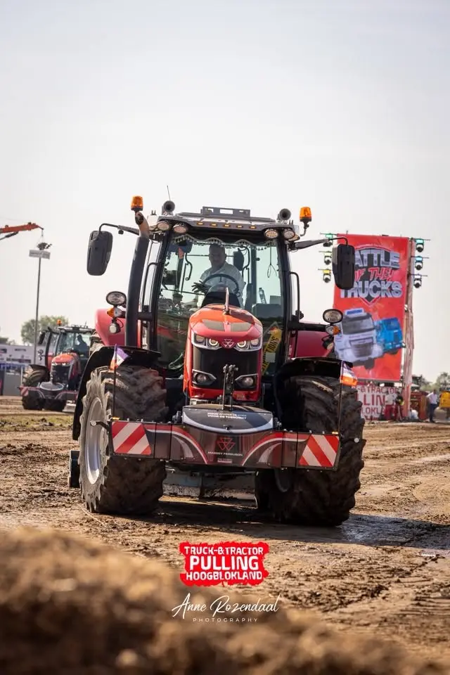 Red tractor with a front weight bumper on a dirt track at an outdoor event with people and banners in the background