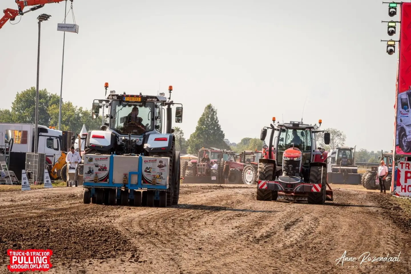 Two tractors on a dirt track, each fitted with large front weight bumpers, with people and vehicles in the background