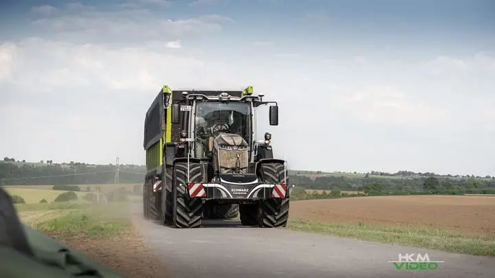 A tractor with a front weight bumper drives on a rural road pulling a large green trailer with fields in the background