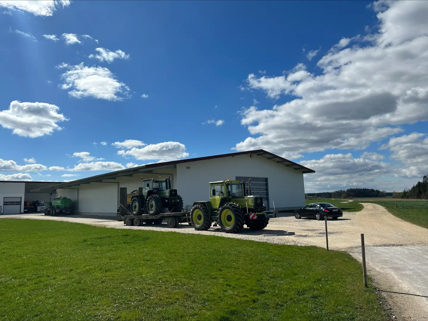 Two green tractors, one with a front weight bumper, are parked outside a large white farm building on a sunny day