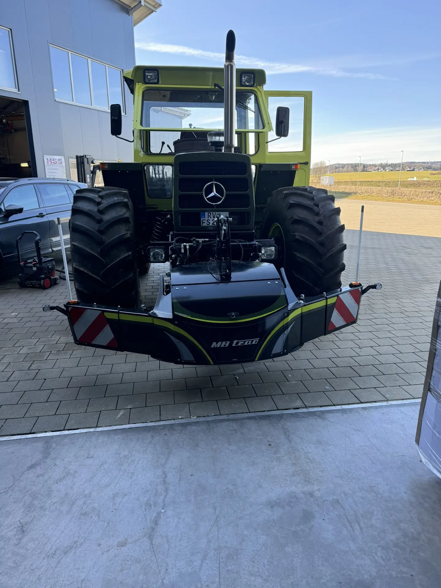 Green tractor with large front tires fitted with an MB trac front weight bumper on a paved area outside a building
