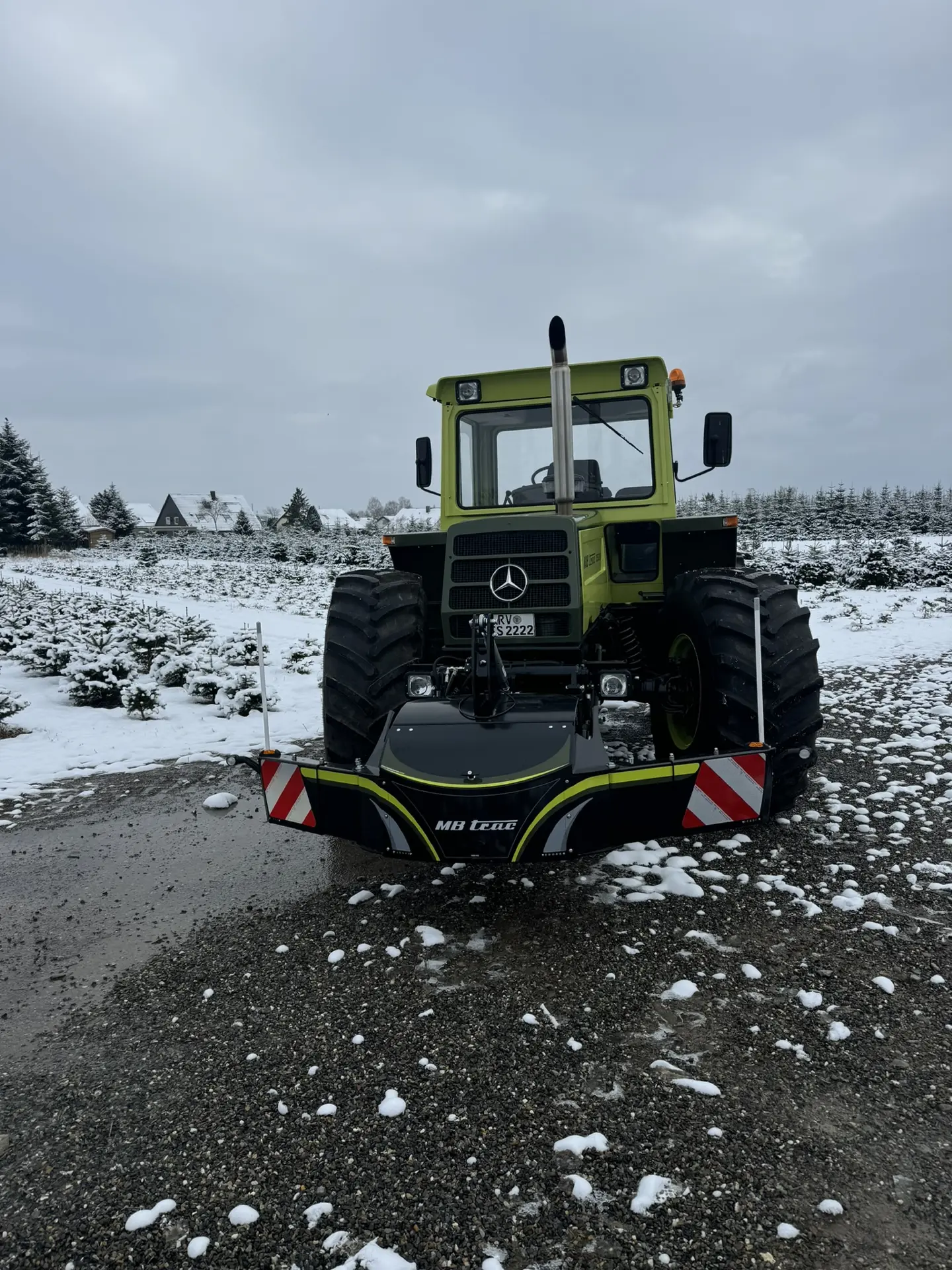 Green tractor with large tires and an MB trac front weight bumper, parked on a snowy gravel surface