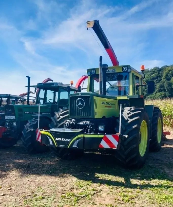 A green and yellow MB trac tractor with a mounted black front weight bumper, parked beside other tractors outdoors