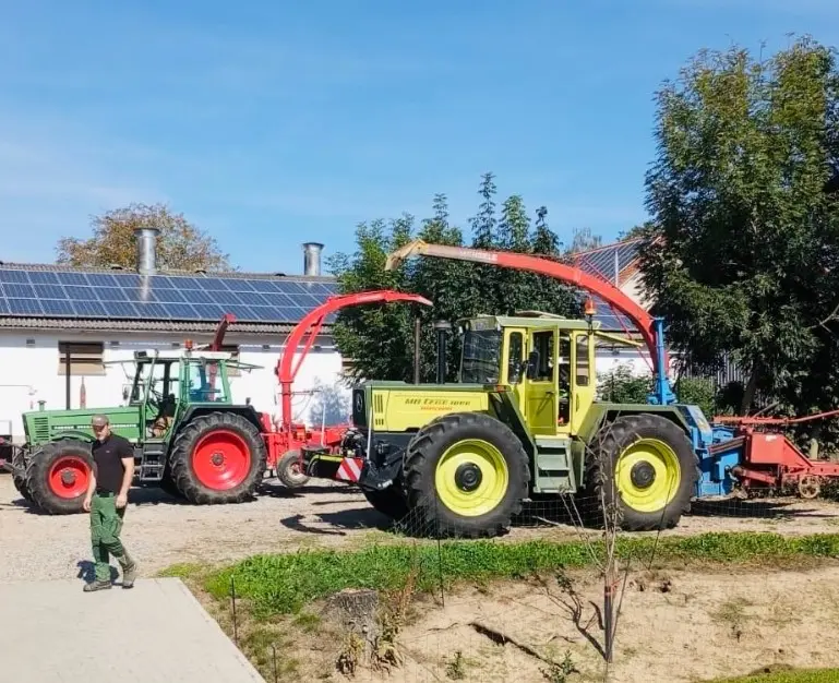 Two tractors with front weight bumpers and attached agricultural machinery are parked outdoors near buildings and trees