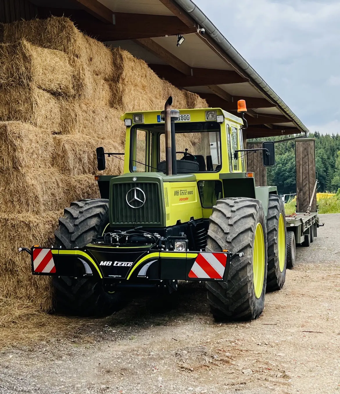 A large tractor with a visible MB trac front weight bumper is parked beside stacked hay bales under a shed roof