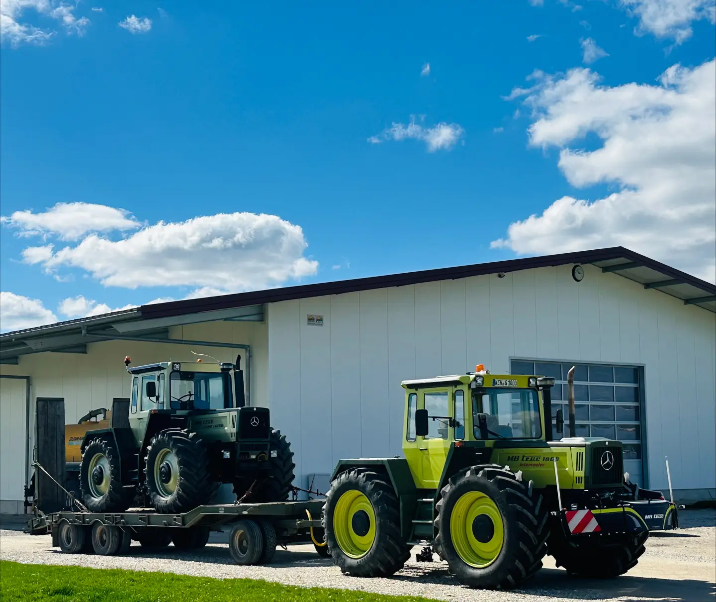 Two green tractors, one on a trailer and one parked in front, both with front weight bumpers, outside a building
