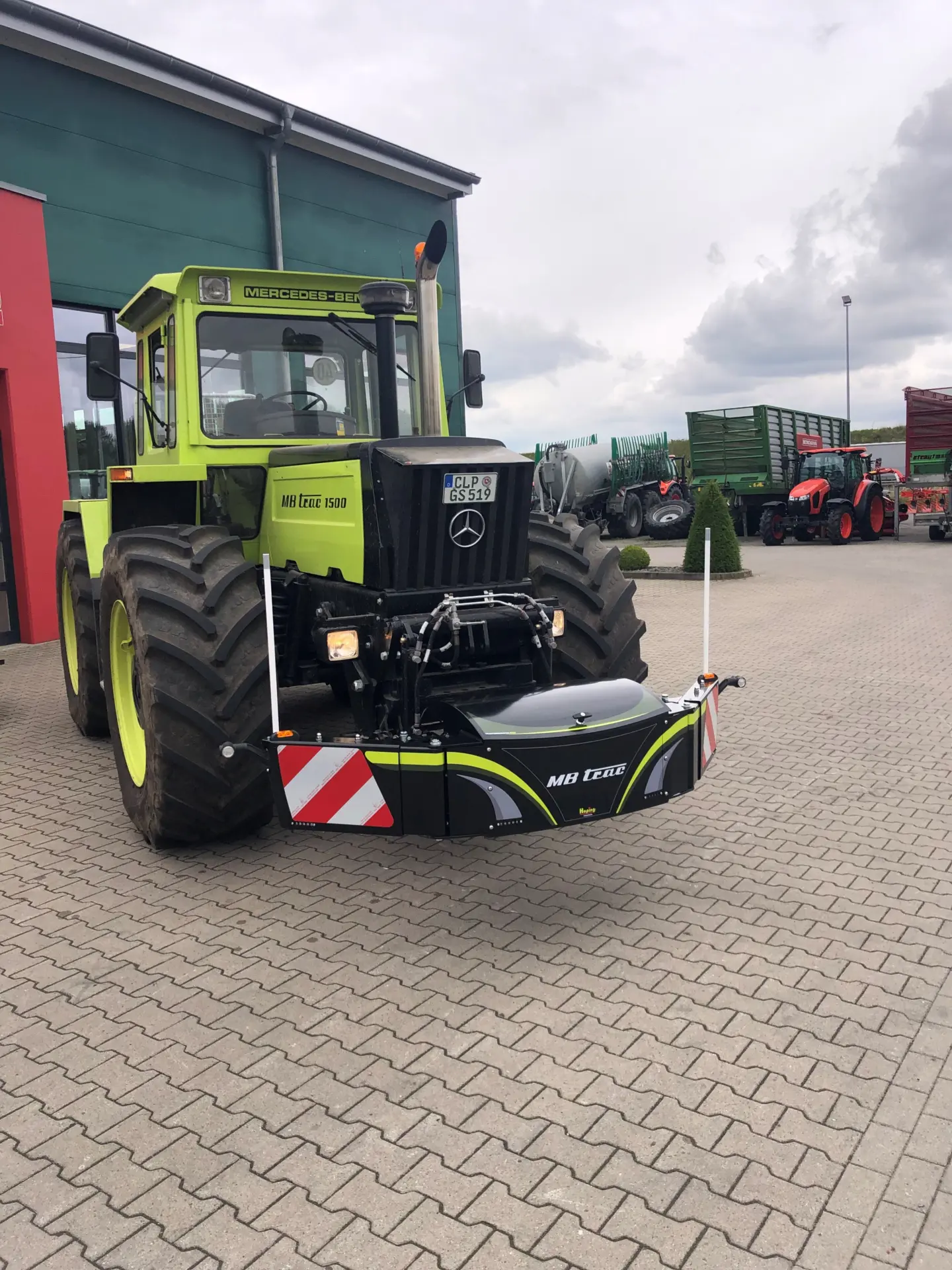 A large green tractor with a black front weight bumper featuring reflective stripes and MB trac branding attached.