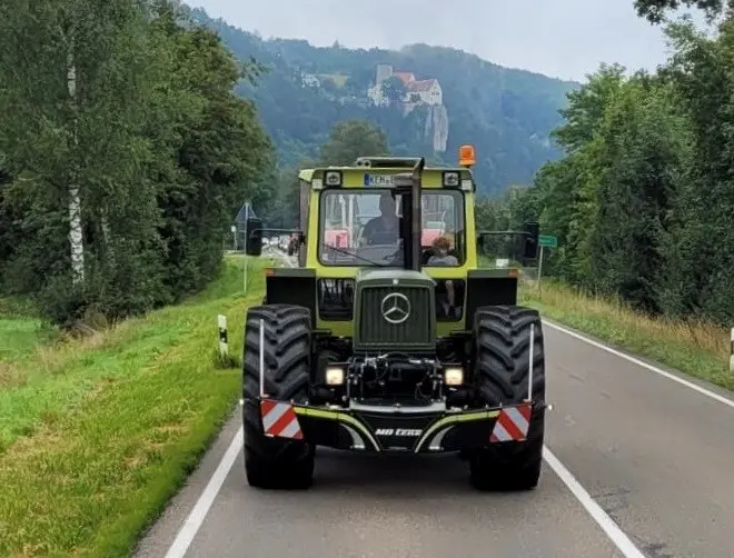 A green tractor with a front weight bumper drives on a rural road with trees and a hilltop building in the background