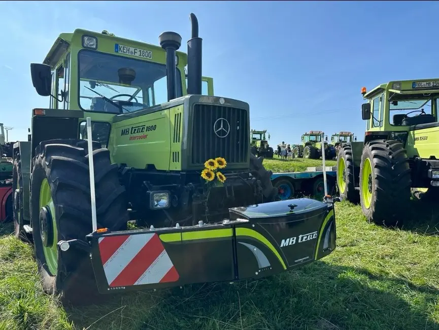 A green tractor with large tires displays a black MB Trac front weight bumper decorated with sunflowers.
