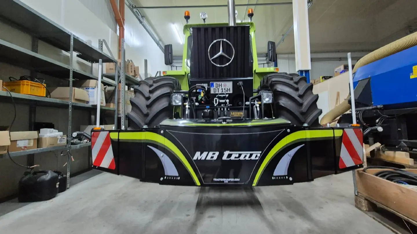 Front view of a tractor indoors with a large black front weight bumper labeled MB trac and red-white warning stripes
