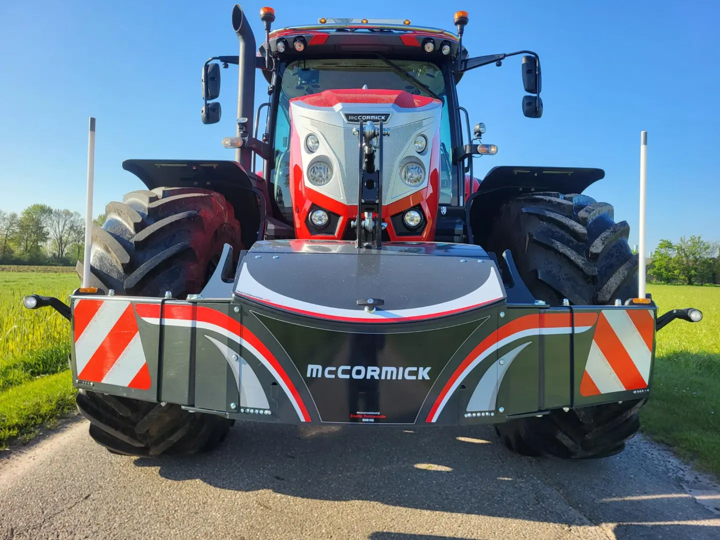 Red tractor with wide tires and a front weight bumper labeled McCormick, parked on a rural road in sunlight