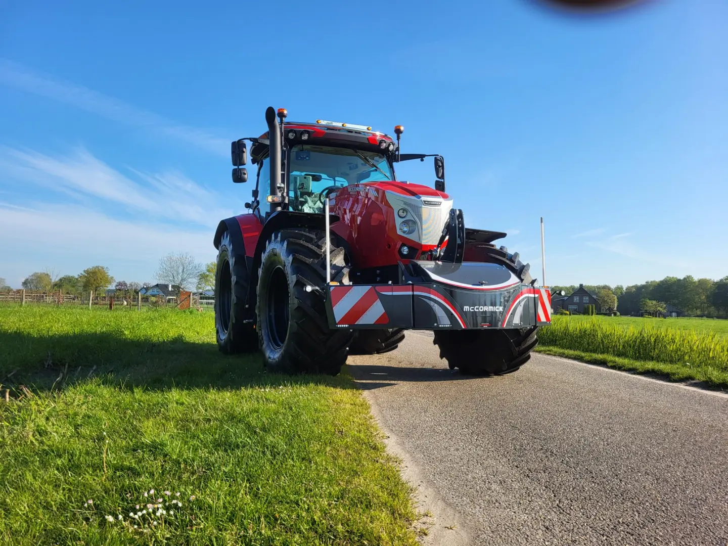 Red tractor with a front weight bumper labeled McCormick parked on a rural road with fields and houses in the background