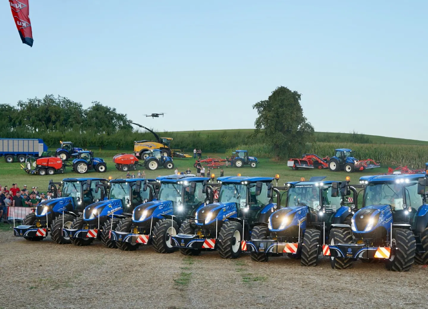 A row of blue tractors each fitted with front weight bumpers, with people and more machinery in the background