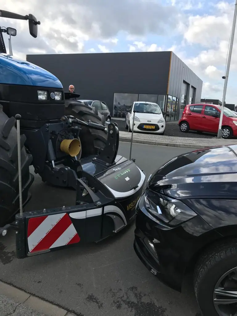 A blue tractor with a front weight bumper is positioned closely in front of a black car on a street.