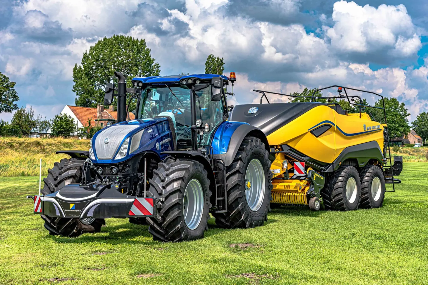 A blue tractor with a front weight bumper is attached to a large yellow trailer on grass with trees in the background