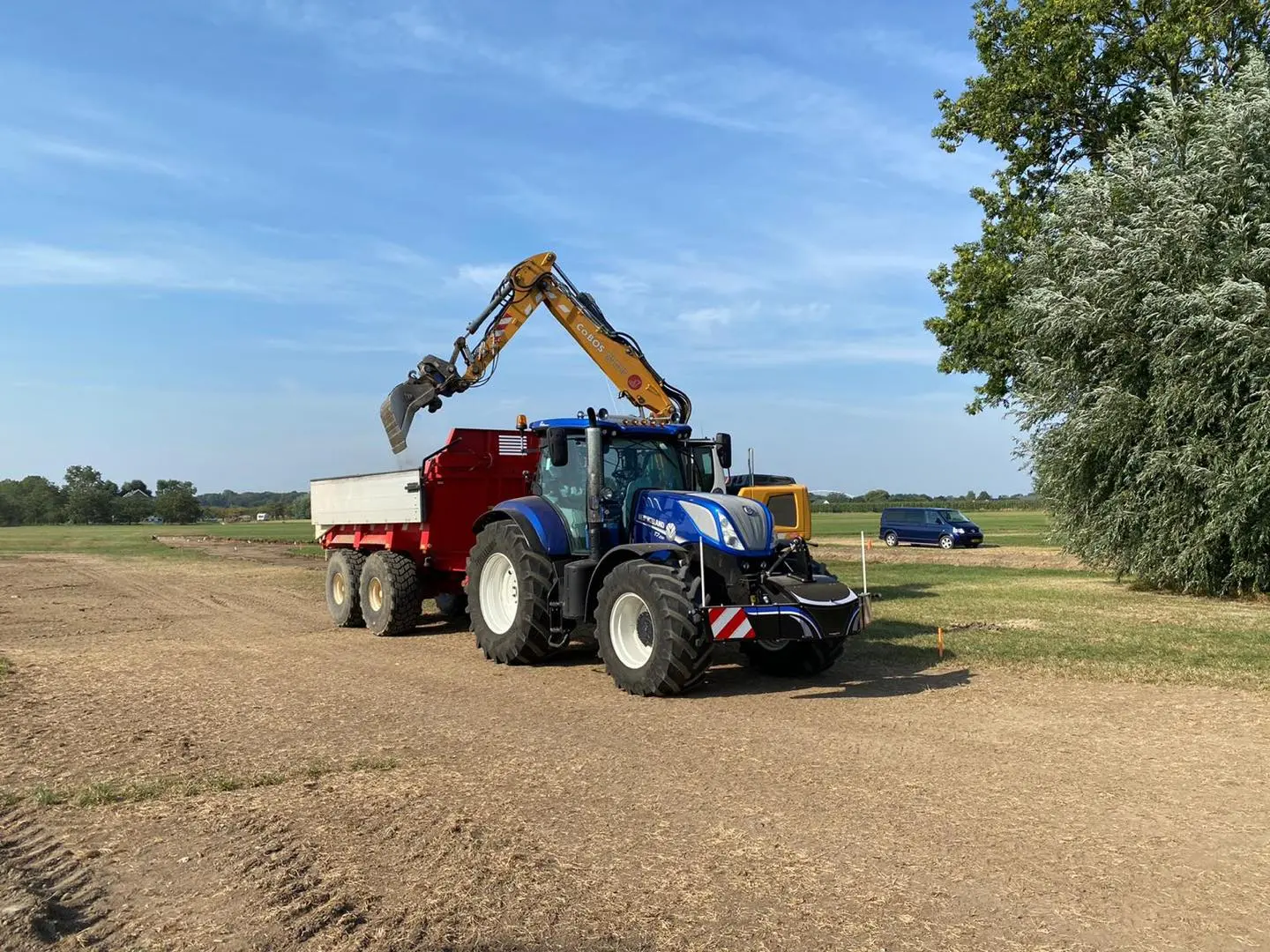 A blue tractor with a front weight bumper attached is pulling a red trailer being loaded by a yellow excavator arm