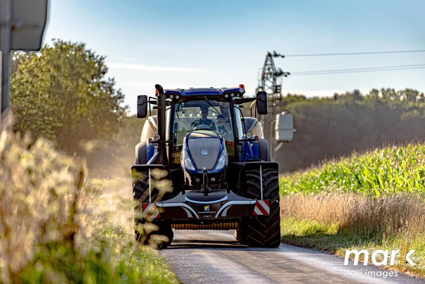 Blue tractor driving on a narrow rural road with a front weight bumper attached, surrounded by green fields