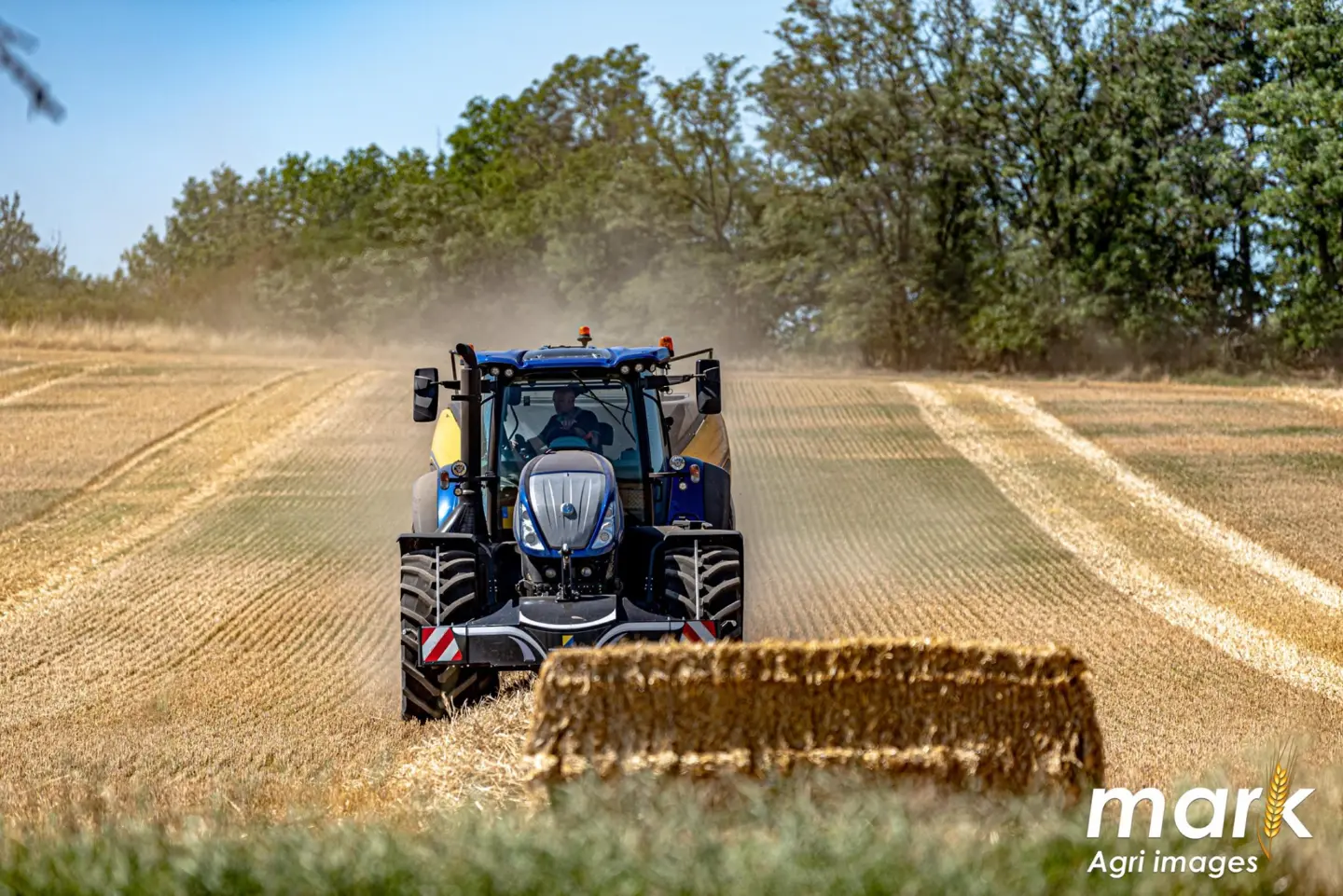A blue tractor with a front weight bumper drives through a harvested field toward a large rectangular straw bale