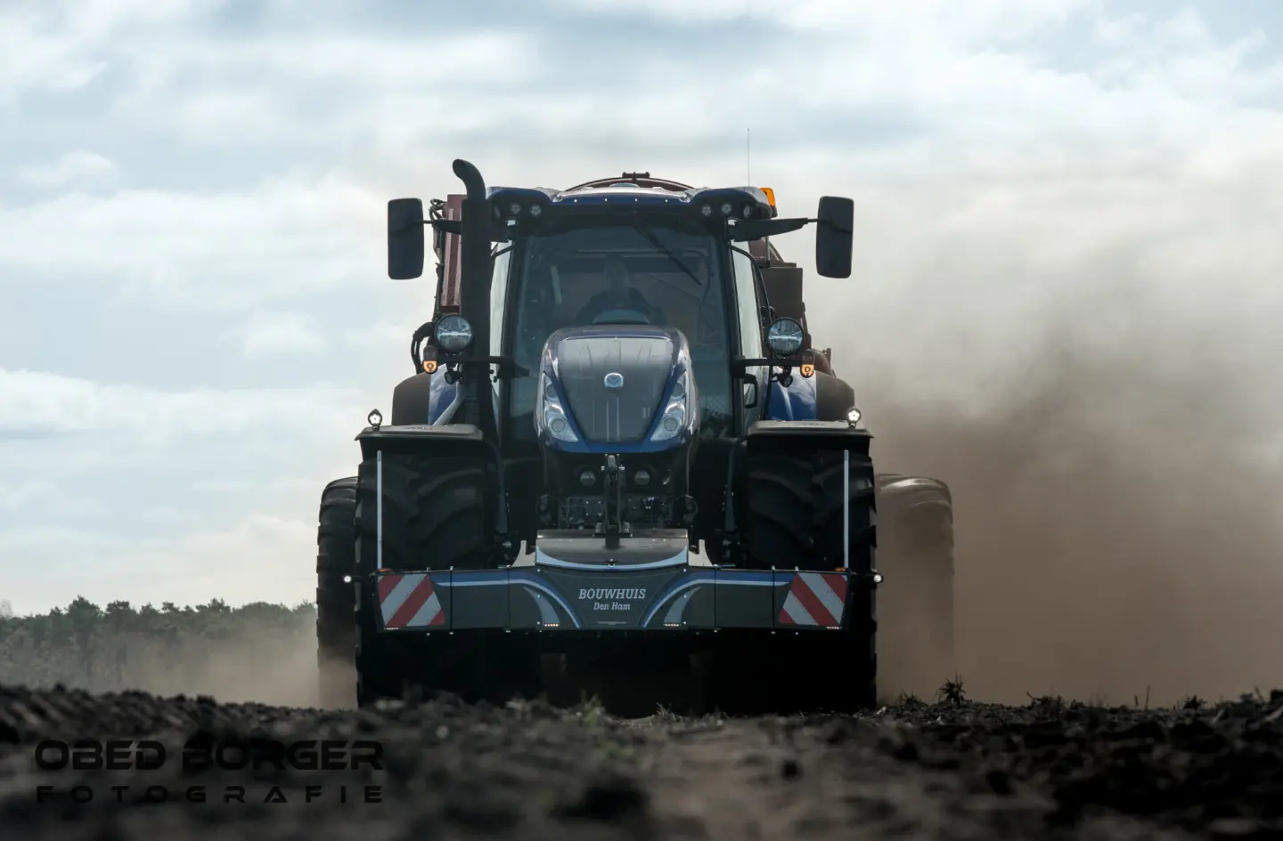 Blue tractor with a front weight bumper drives across a field, kicking up dust behind it under a cloudy sky