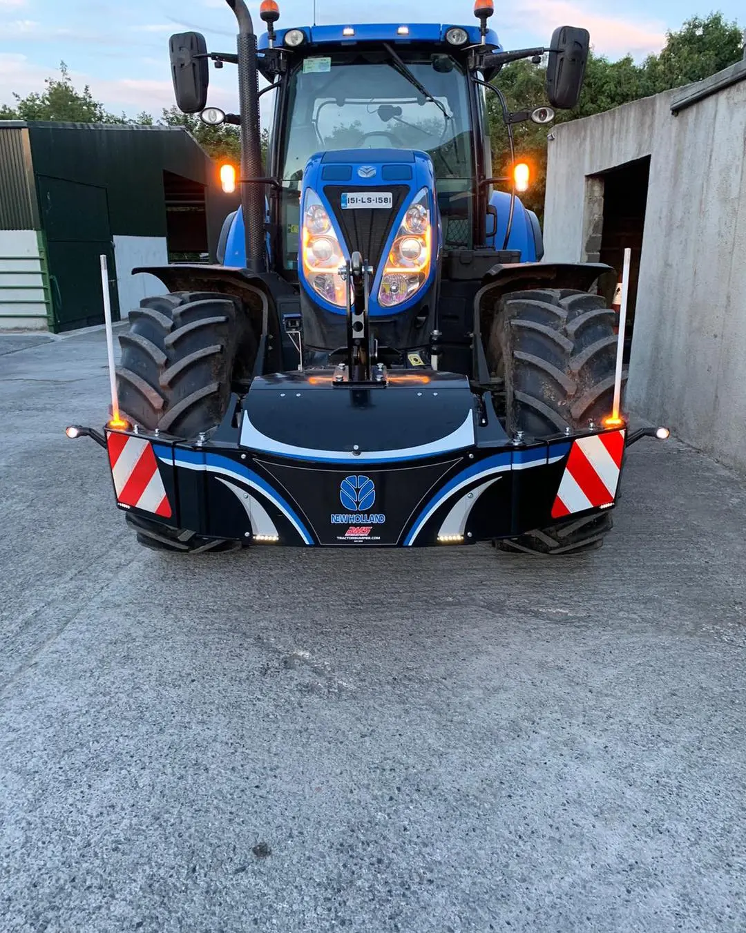 Blue tractor with a front weight bumper featuring New Holland branding, warning stripes and lights on a concrete yard