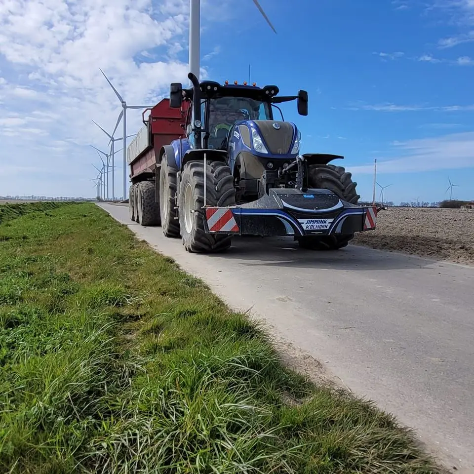 Blue tractor with a front weight bumper on a rural road, pulling a red trailer with wind turbines in the background