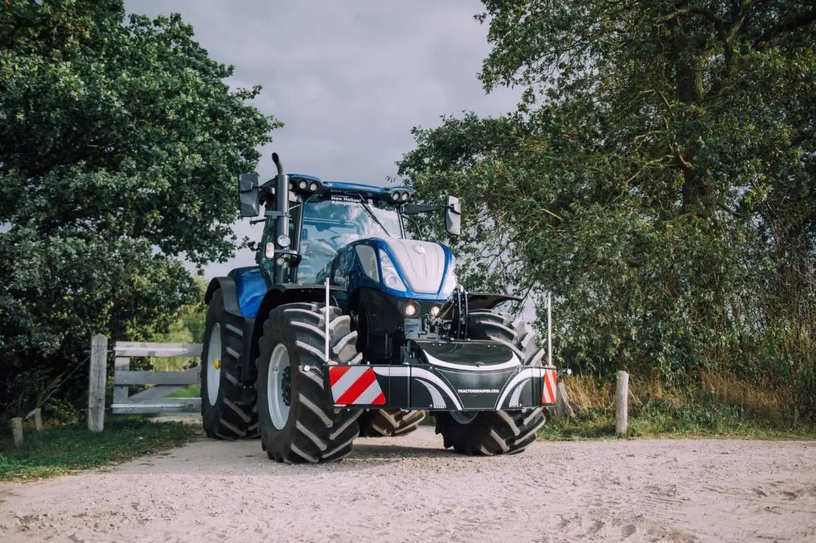 A blue tractor fitted with a black front weight bumper featuring red and white reflective safety markings.