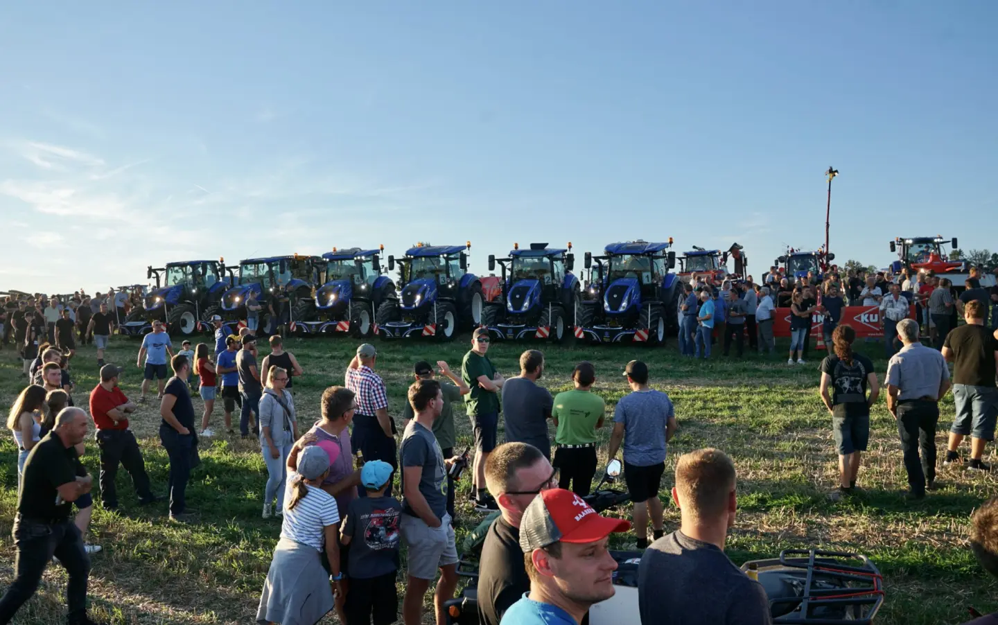 Several blue tractors fitted with front weight bumpers lined up in a field with a crowd of people gathered around