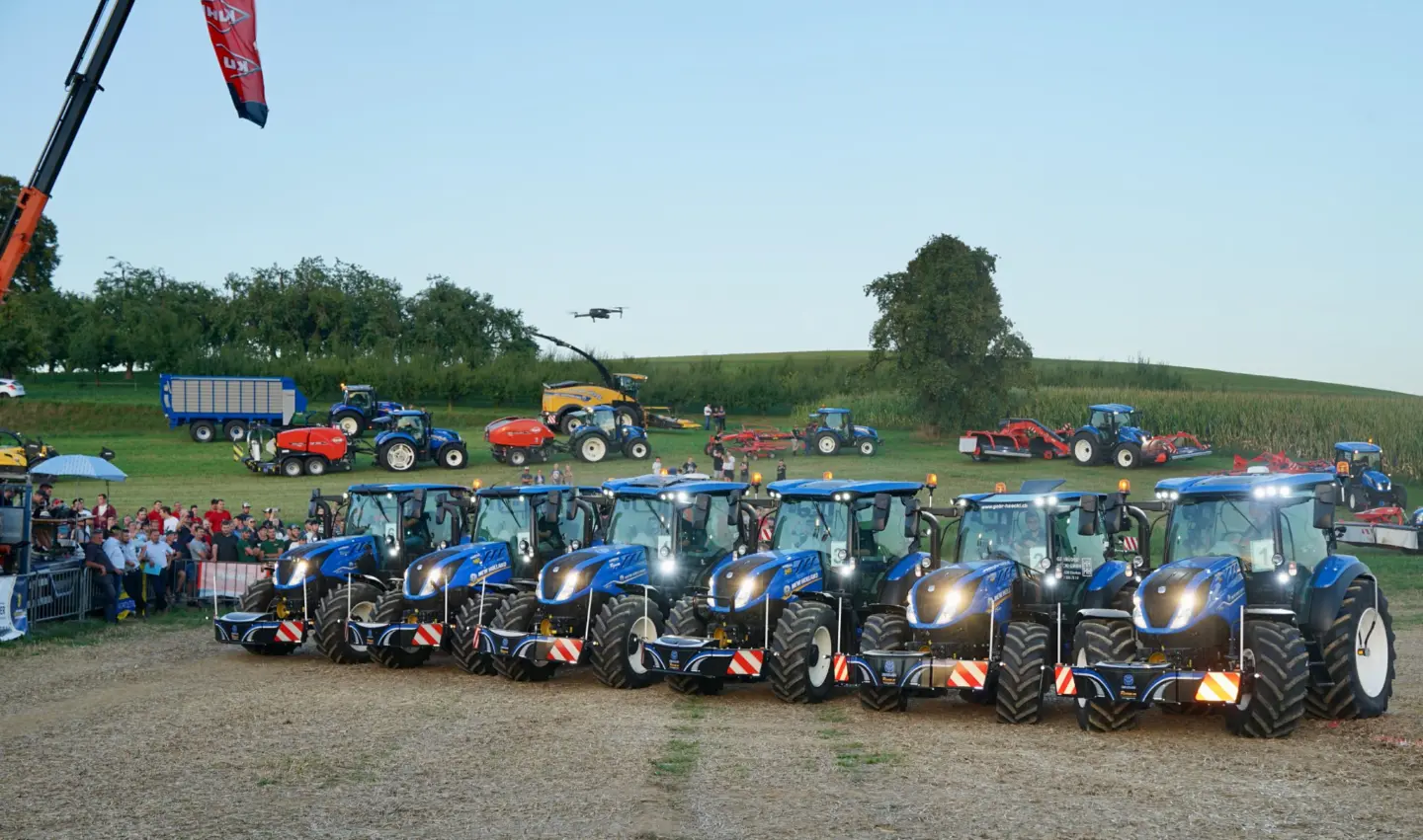 Seven blue tractors equipped with front weight bumpers are parked in a row at an outdoor event with spectators.