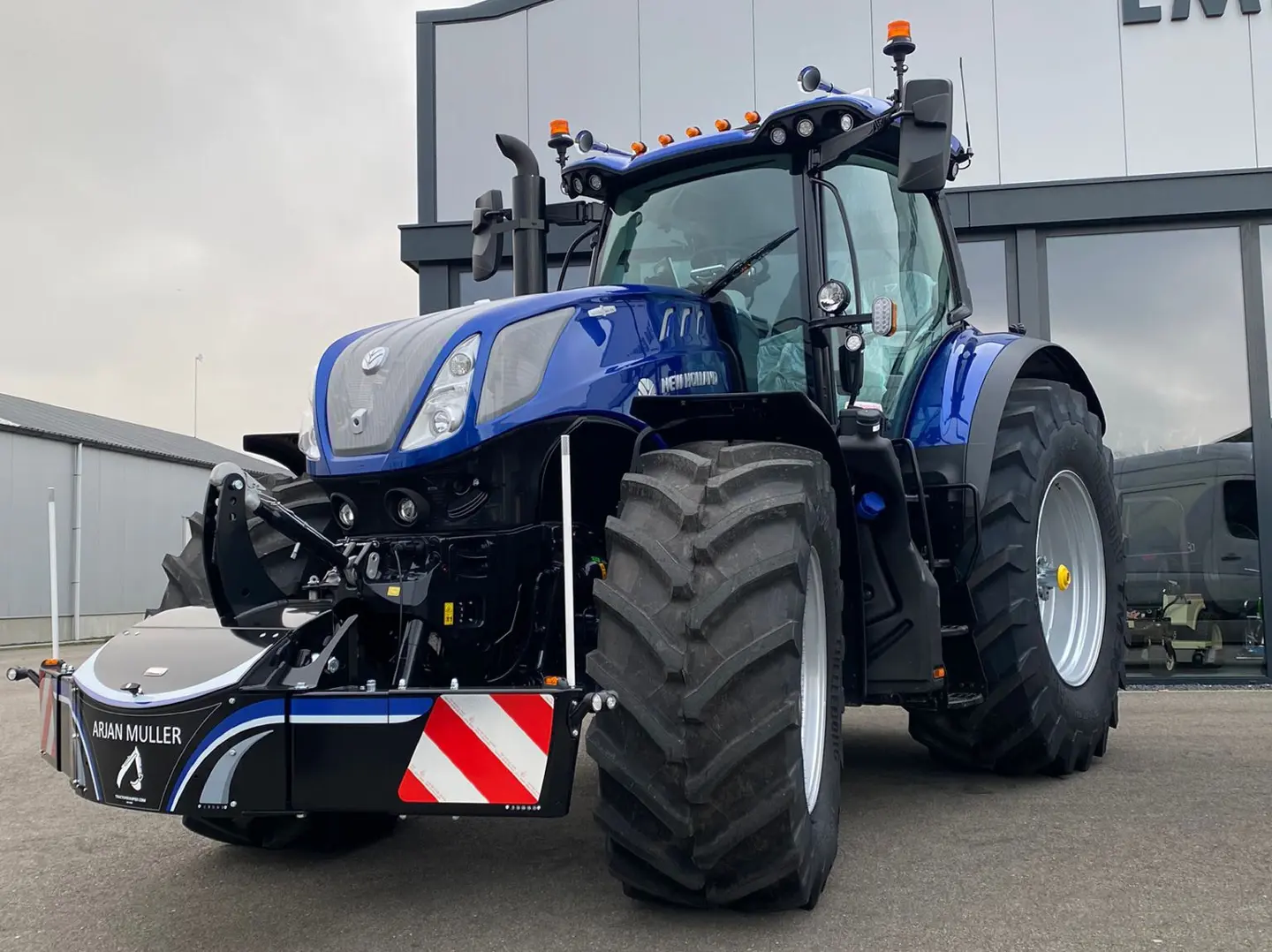 A blue tractor fitted with a large black front weight bumper that has warning stripes and marker poles attached