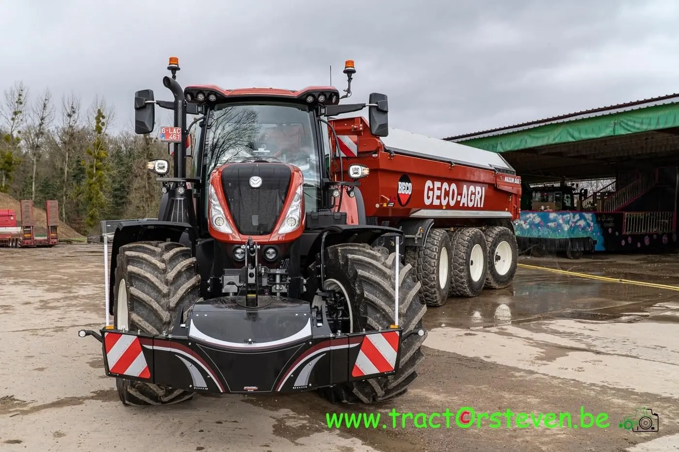 Large tractor with a front-mounted weight bumper and reflective warning panels, hitched to a red agricultural trailer